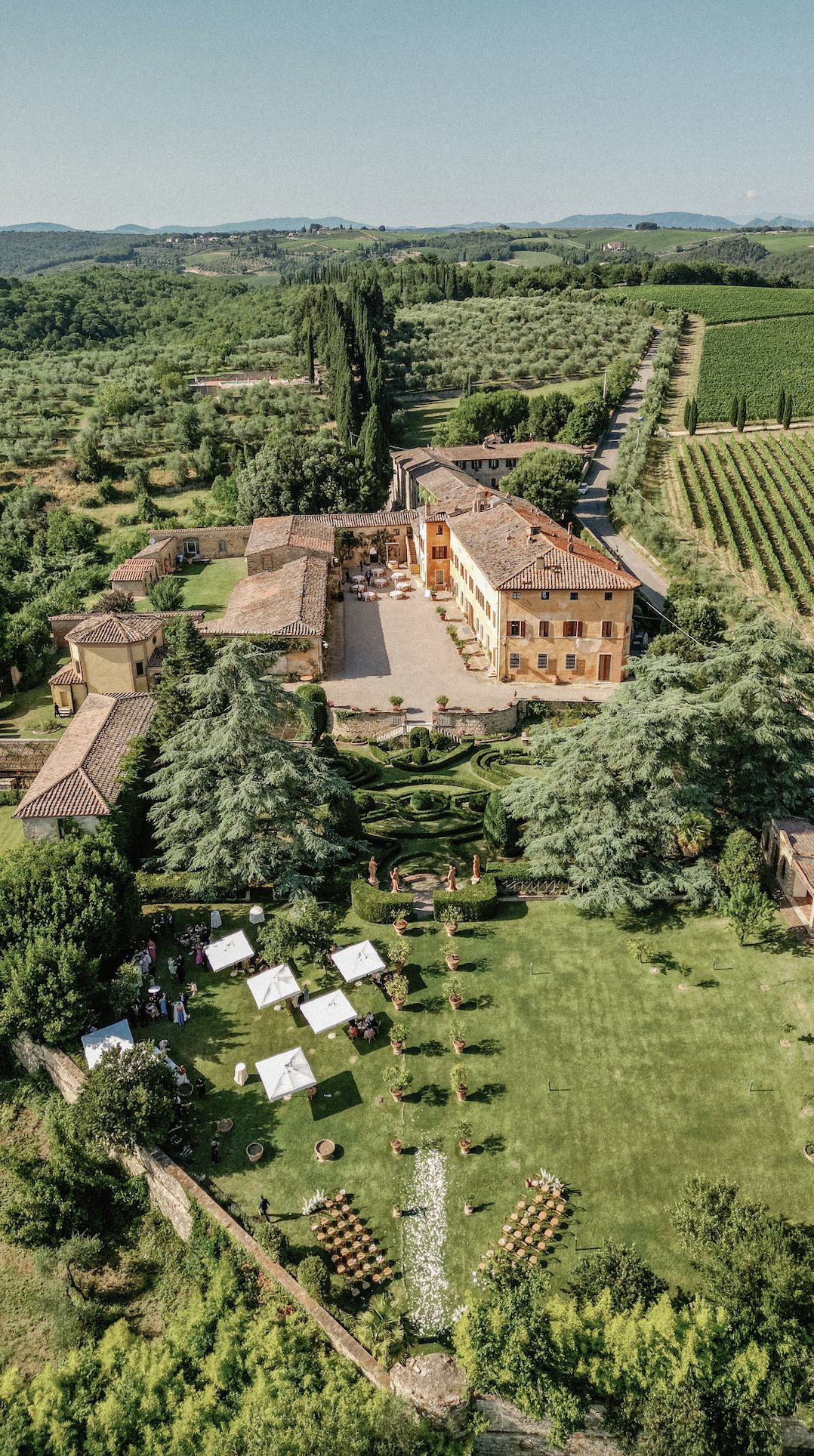Aerial view of a historic Italian villa estate with formal gardens and a lawn set for a wedding ceremony, surrounded by olive groves and rolling hills